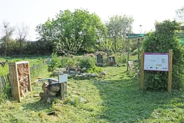 Jardin enherbé cloturé avec de la ganivelle contenant des abris à insecte, une rocaille, des arbustes, un panneau informatif et une ombrière