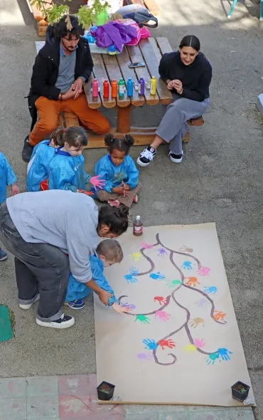des enfants, accompagnés d'adultes posent leur main pleine de peinture sur une fresque en forme d'arbre pour former des feuilles de mains