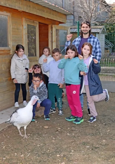 un groupe d'enfants et leurs animateurs sont devant un poulailler et derrière une dinde en train de faire un signe avec leurs doigts