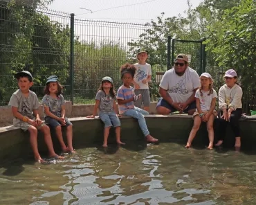 Un groupe d'enfants et leur animateur sont assis autour de la mare des canards, les pieds dans l'eau
