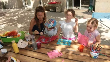 Une animatrice colle avec un enfant un éléments naturel et deux autres enfants nous montrent leurs œuvres