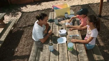 Une animatrice et deux enfants assis à une table mettent des plantes à sécher pour leur herbier