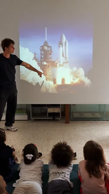 Un groupe d'enfants est allogé et un animateur montre sur grand écran la photo du décollage d'une navette spatiale