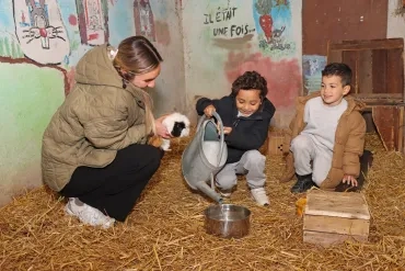 deux enfants et leur animatrice sont dans un enclos. Un enfant verse de l'eau dans une gamelle et l'animatrice tient un cochon d'Inde