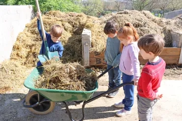 Un groupe d'enfants tient une brouette et l'un d'eux met de la paille à l'intérieur à l'aide d'une pelle