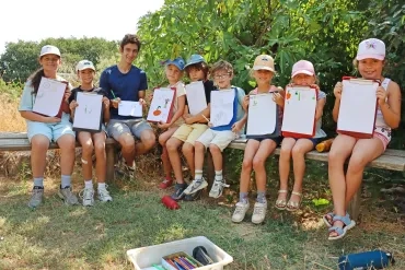 Un groupe d'enfants et leur animateur sont assis sur des bancs et montrent des dessins