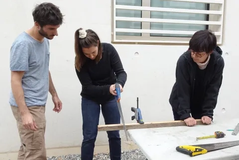 Une adulte utilise une scie à bois pour couper un tasseau maintenu par une autre adulte. Un animateur supervise.