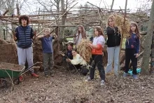 un groupe d'enfants et leurs animateurs montrent de la paille et de la boue devant une cabane